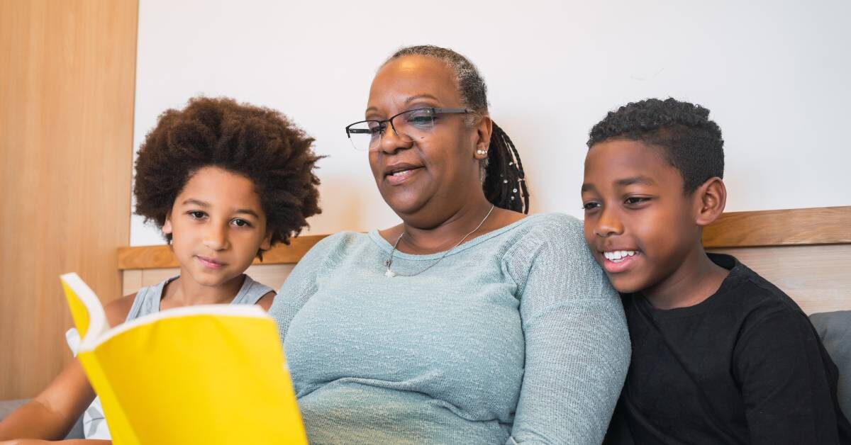 A black grandmother reads a children's book to her two black grandchildren. Her grandchildren are listening intently.