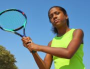 A teenage girl in a neon yellow shirt against a bright blue sky, holding a colorful tennis racquet and warming up her swing.