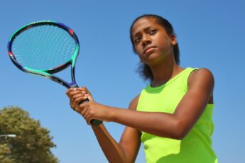 A teenage girl in a neon yellow shirt against a bright blue sky, holding a colorful tennis racquet and warming up her swing.