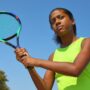 A teenage girl in a neon yellow shirt against a bright blue sky, holding a colorful tennis racquet and warming up her swing.