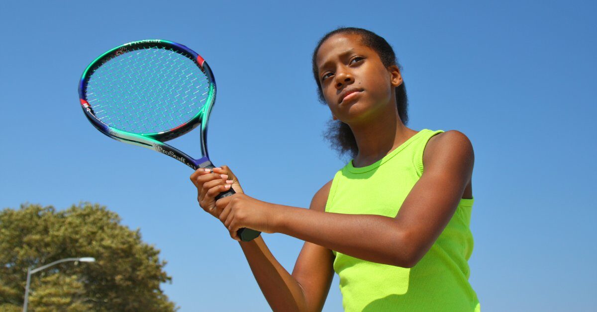 A teenage girl in a neon yellow shirt against a bright blue sky, holding a colorful tennis racquet and warming up her swing.