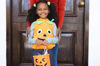 A young girl wearing a pumpkin vest smiling and holding a plastic pumpkin full of candy. Her mother stands behind her.