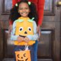 A young girl wearing a pumpkin vest smiling and holding a plastic pumpkin full of candy. Her mother stands behind her.