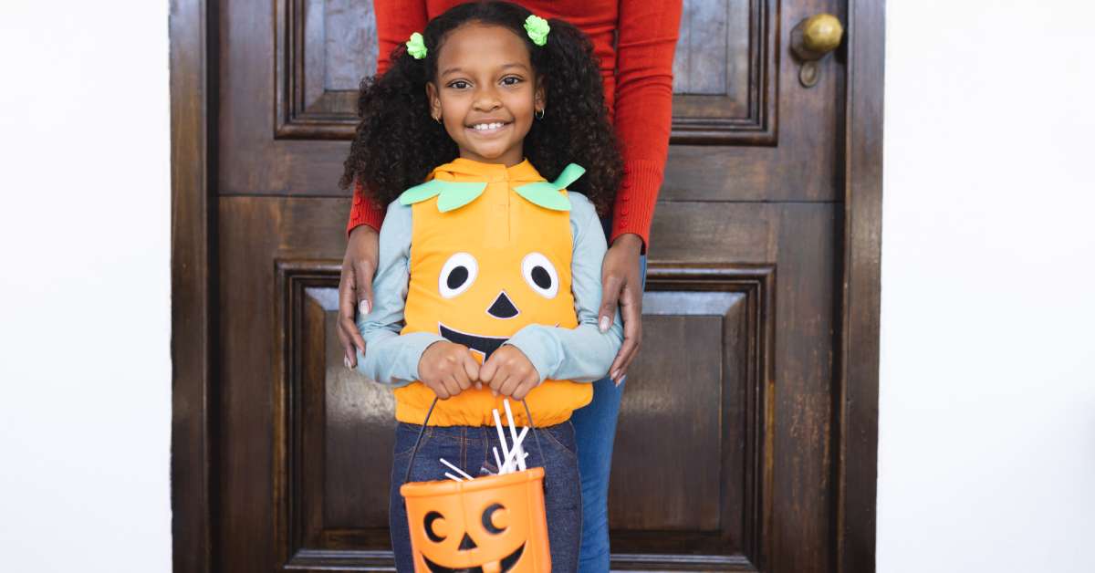 A young girl wearing a pumpkin vest smiling and holding a plastic pumpkin full of candy. Her mother stands behind her.