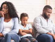 A Black mother and father sitting on a couch with their daughter; the young girl appears upset with her arms crossed.