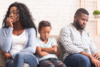 A Black mother and father sitting on a couch with their daughter; the young girl appears upset with her arms crossed.