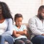 A Black mother and father sitting on a couch with their daughter; the young girl appears upset with her arms crossed.