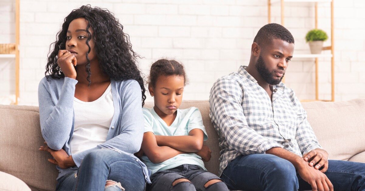 A Black mother and father sitting on a couch with their daughter; the young girl appears upset with her arms crossed.