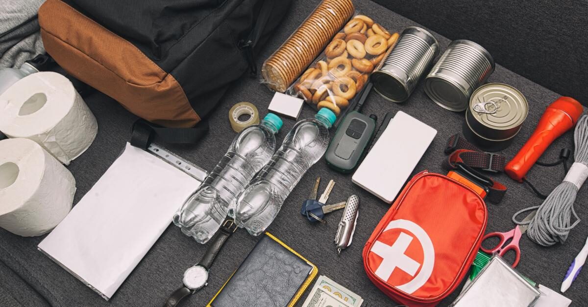 A black backpack and emergency supplies like water bottles, money, and a first aid kit on a couch.