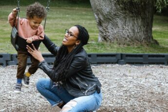 A young mother kneeling down beside her child, who is sitting in a toddler swing, and helping him swing.