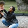 A young mother kneeling down beside her child, who is sitting in a toddler swing, and helping him swing.