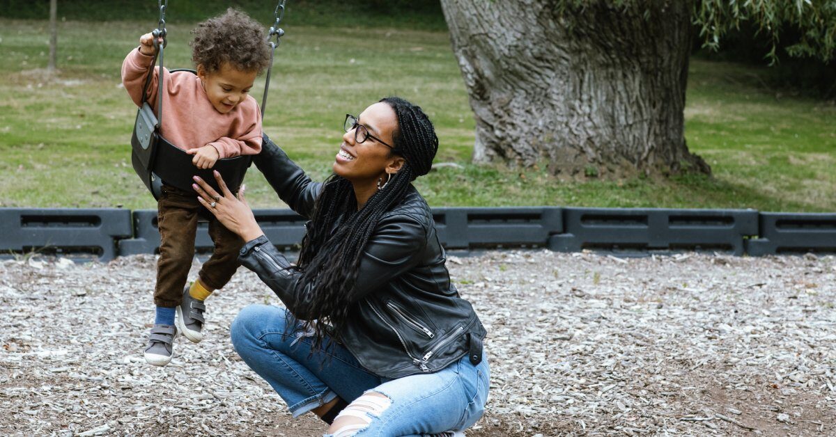 A young mother kneeling down beside her child, who is sitting in a toddler swing, and helping him swing.