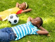 Two young Black children laughing and playing together in the yard. They have a soccer ball close by and smiling faces.