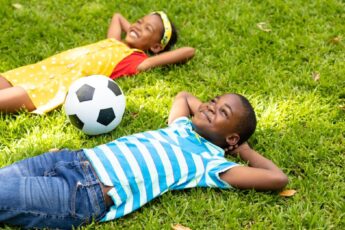 Two young Black children laughing and playing together in the yard. They have a soccer ball close by and smiling faces.