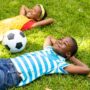 Two young Black children laughing and playing together in the yard. They have a soccer ball close by and smiling faces.