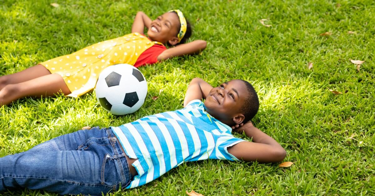 Two young Black children laughing and playing together in the yard. They have a soccer ball close by and smiling faces.