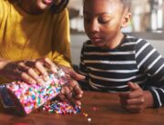 A mother and her child sit at a table in their kitchen. The mother pours beads onto the table while her child watches.