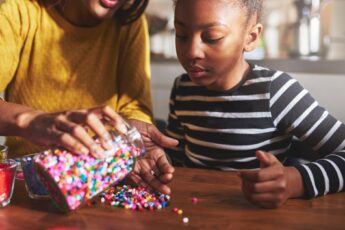 A mother and her child sit at a table in their kitchen. The mother pours beads onto the table while her child watches.