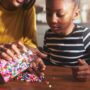 A mother and her child sit at a table in their kitchen. The mother pours beads onto the table while her child watches.