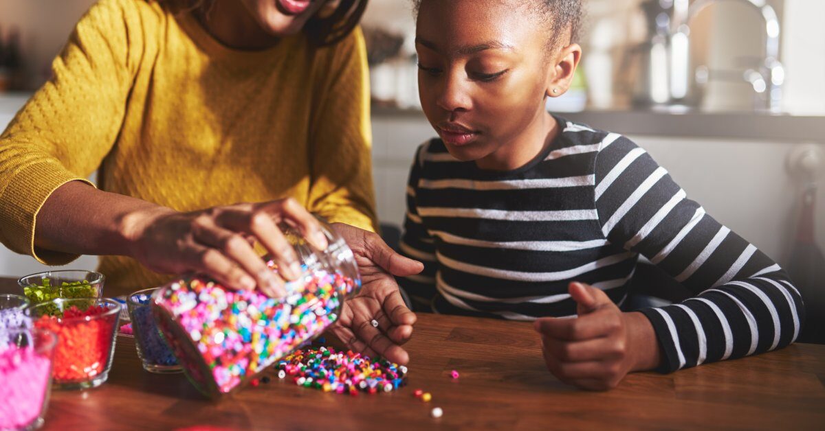 A mother and her child sit at a table in their kitchen. The mother pours beads onto the table while her child watches.