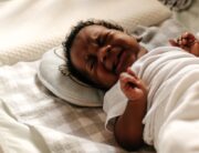 A little Black baby crying in their crib. They are wrapped in a white blanket and have their fists clutched.