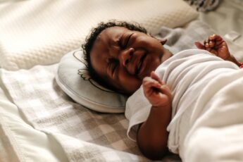 A little Black baby crying in their crib. They are wrapped in a white blanket and have their fists clutched.