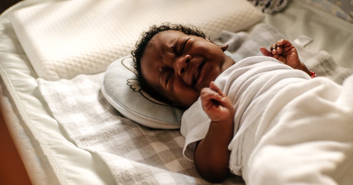 A little Black baby crying in their crib. They are wrapped in a white blanket and have their fists clutched.