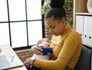 A young black mother sitting at a desk, looking down, and holding her infant to her chest as they breastfeed.