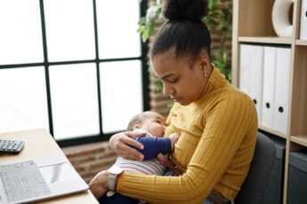 A young black mother sitting at a desk, looking down, and holding her infant to her chest as they breastfeed.