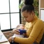 A young black mother sitting at a desk, looking down, and holding her infant to her chest as they breastfeed.