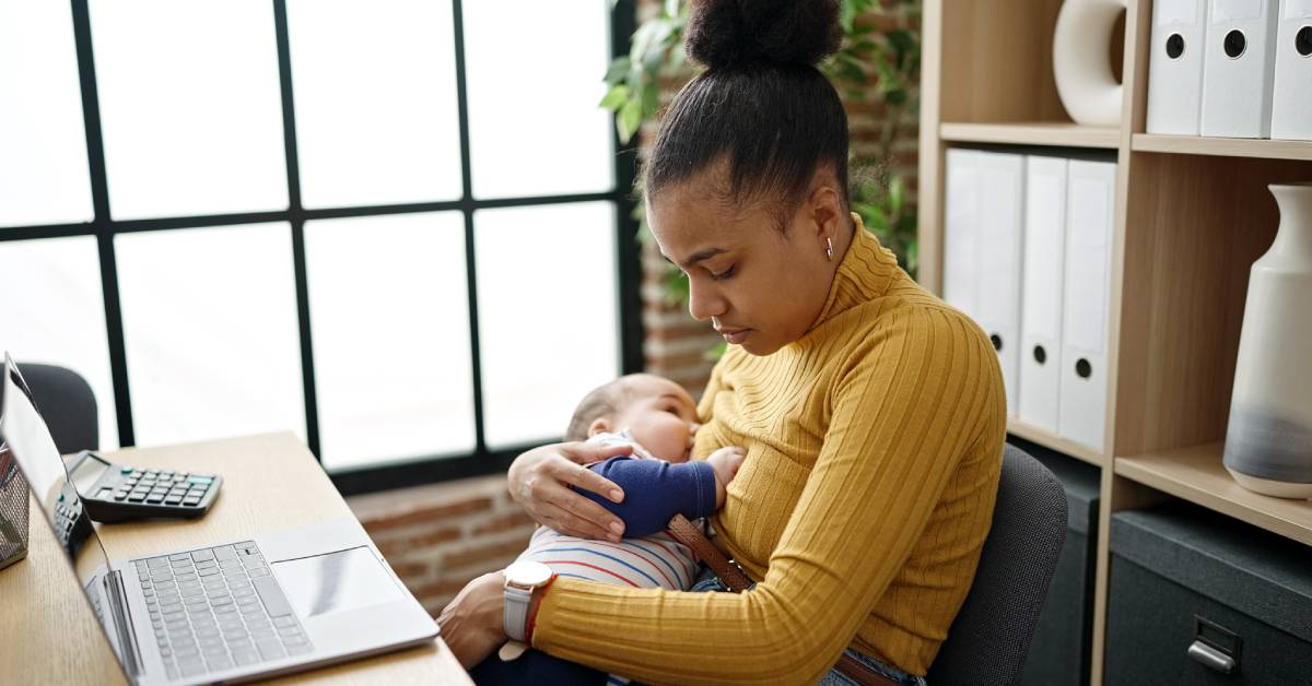 A young black mother sitting at a desk, looking down, and holding her infant to her chest as they breastfeed.