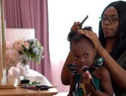 A mother combing through her daughter’s coily hair. They sit facing a vanity set with her daughter holding a makeup brush.