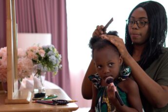 A mother combing through her daughter’s coily hair. They sit facing a vanity set with her daughter holding a makeup brush.