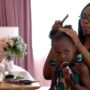 A mother combing through her daughter’s coily hair. They sit facing a vanity set with her daughter holding a makeup brush.