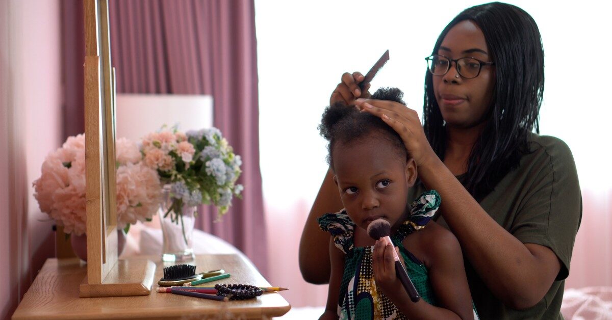 A mother combing through her daughter’s coily hair. They sit facing a vanity set with her daughter holding a makeup brush.