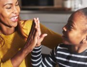 A Black mother in a yellow shirt grinning as she high-fives her young daughter sporting a striped shirt and bun.