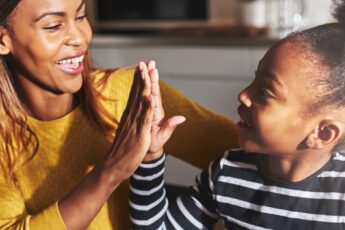 A Black mother in a yellow shirt grinning as she high-fives her young daughter sporting a striped shirt and bun.