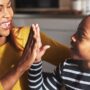 A Black mother in a yellow shirt grinning as she high-fives her young daughter sporting a striped shirt and bun.