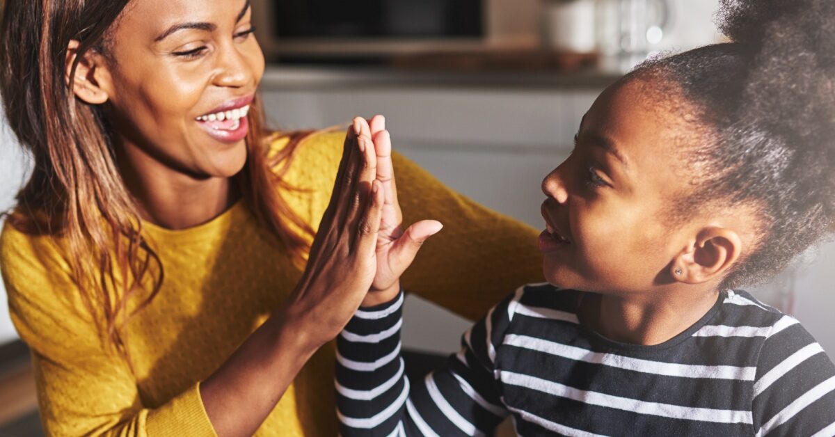 A Black mother in a yellow shirt grinning as she high-fives her young daughter sporting a striped shirt and bun.