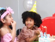 Three girls in birthday hats gather around a table. The birthday girl leans toward a cake while looking at her friend.