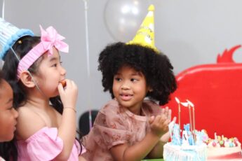 Three girls in birthday hats gather around a table. The birthday girl leans toward a cake while looking at her friend.