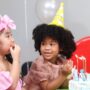 Three girls in birthday hats gather around a table. The birthday girl leans toward a cake while looking at her friend.