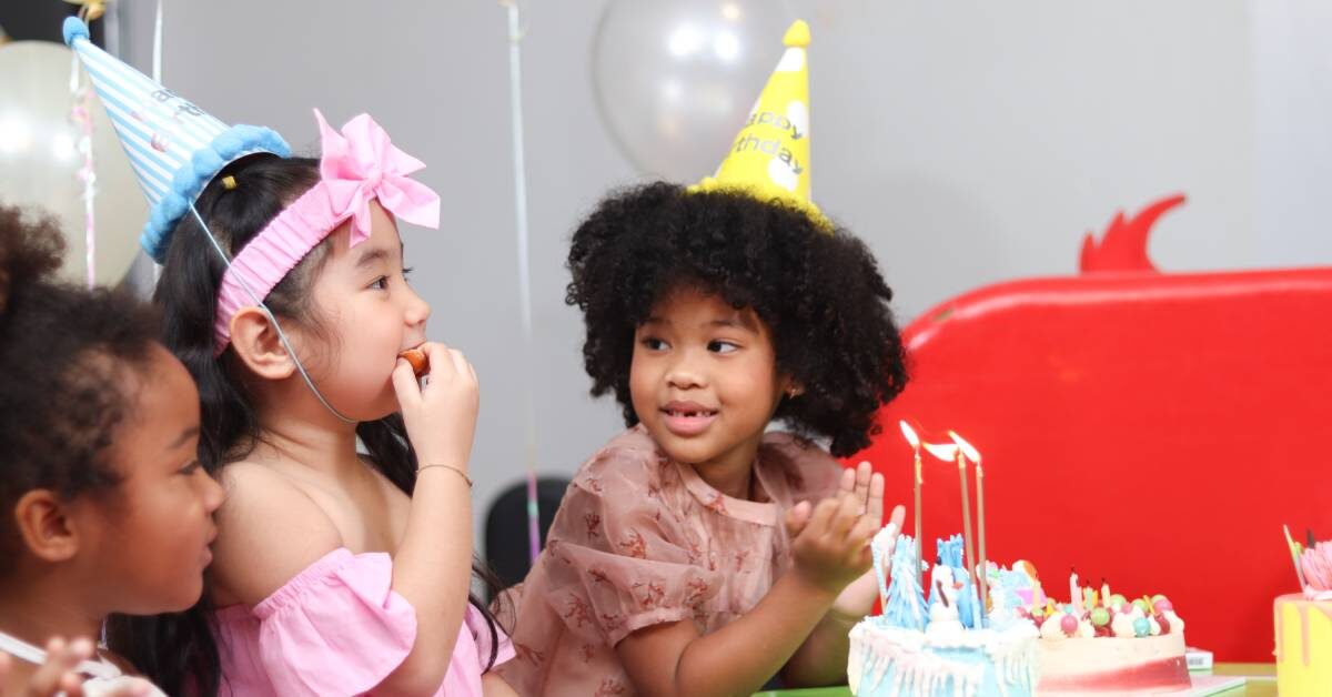 Three girls in birthday hats gather around a table. The birthday girl leans toward a cake while looking at her friend.