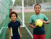 Two young Black children playing with a soccer ball on the field. They're smiling as they stand under the goal net.