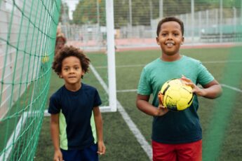 Two young Black children playing with a soccer ball on the field. They're smiling as they stand under the goal net.