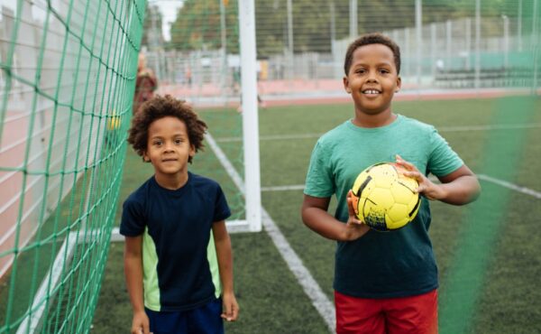 Two young Black children playing with a soccer ball on the field. They're smiling as they stand under the goal net.