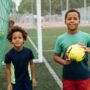 Two young Black children playing with a soccer ball on the field. They're smiling as they stand under the goal net.