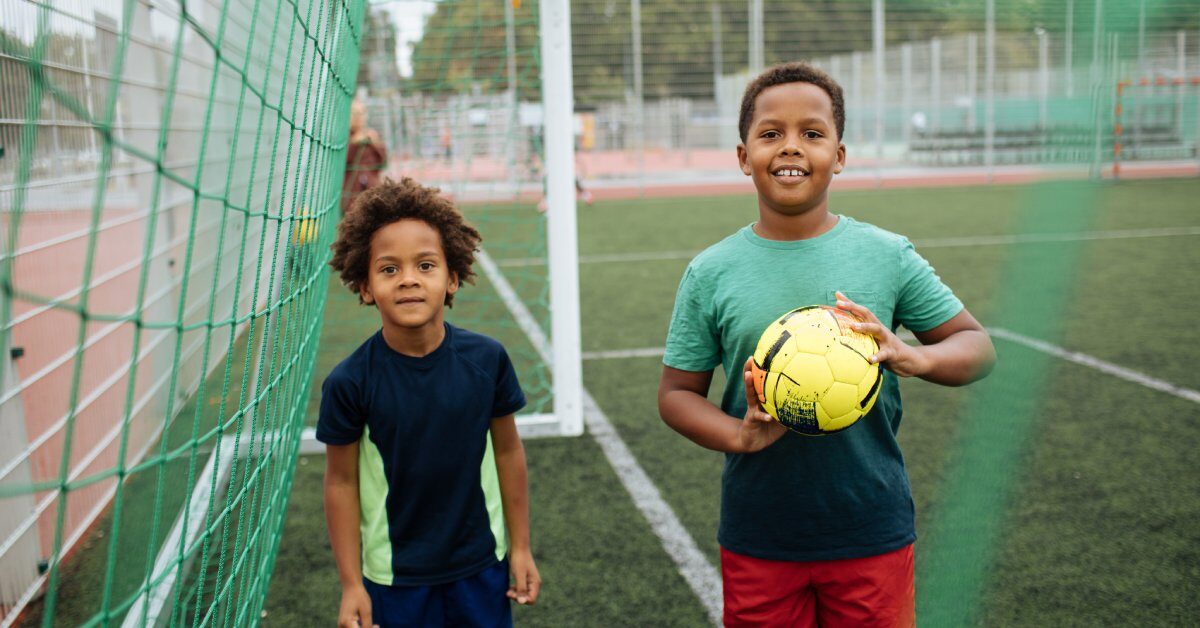 Two young Black children playing with a soccer ball on the field. They're smiling as they stand under the goal net.