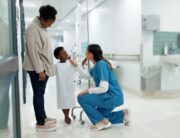 A young Black boy in a hospital gown stands beside his mother while interacting with a healthcare professional in scrubs.