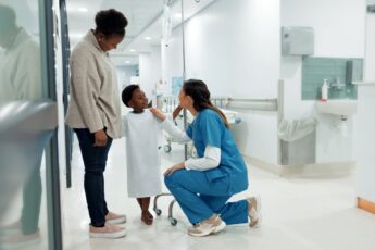 A young Black boy in a hospital gown stands beside his mother while interacting with a healthcare professional in scrubs.